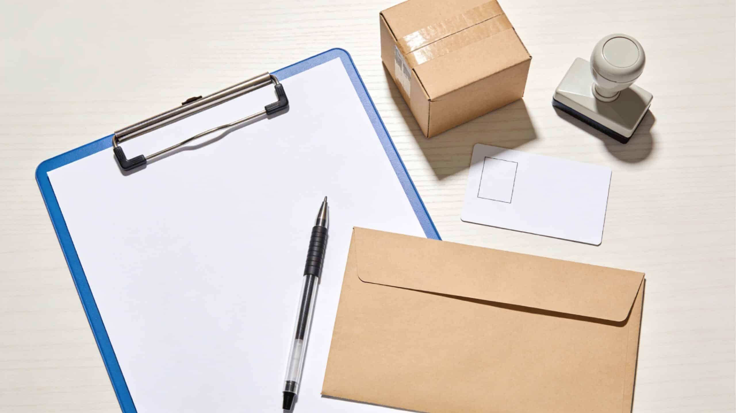 Bureau clair avec enveloppe kraft, petit colis carton, feuille blanche sur clipboard, stylo, carte d’identité retournée et tampon encreur neutre.